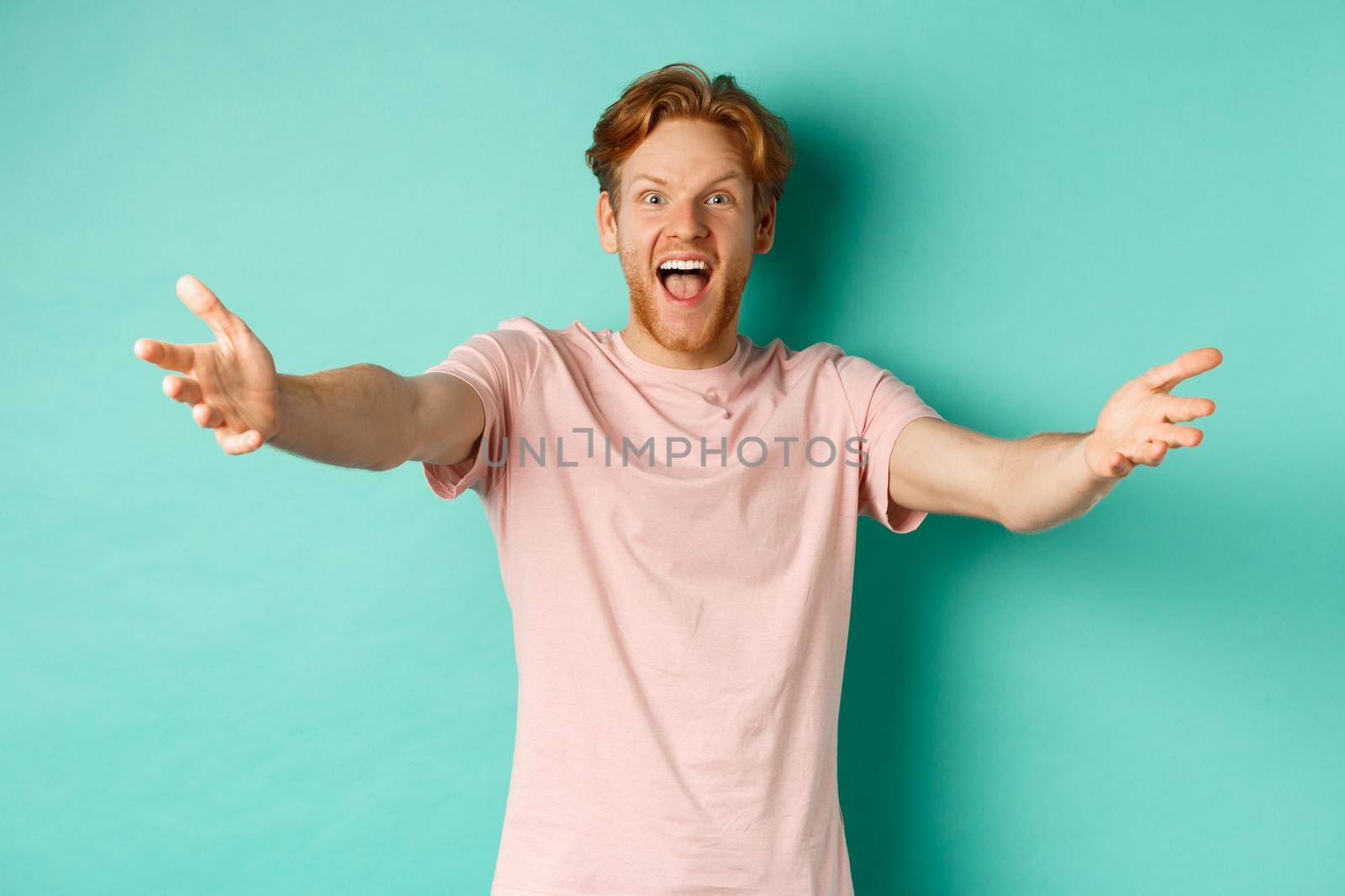 Friendly and happy young man with ginger hair, stretch out hands in warm welcome, reaching for hug and smiling joyfully, standing in t-shirt over mint background by Benzoix