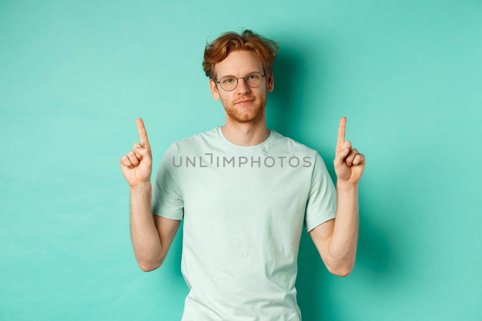 Skeptical and unamused redhead man in glasses and t-shirt, pointing fingers up and showing something useless, standing against mint background by Benzoix