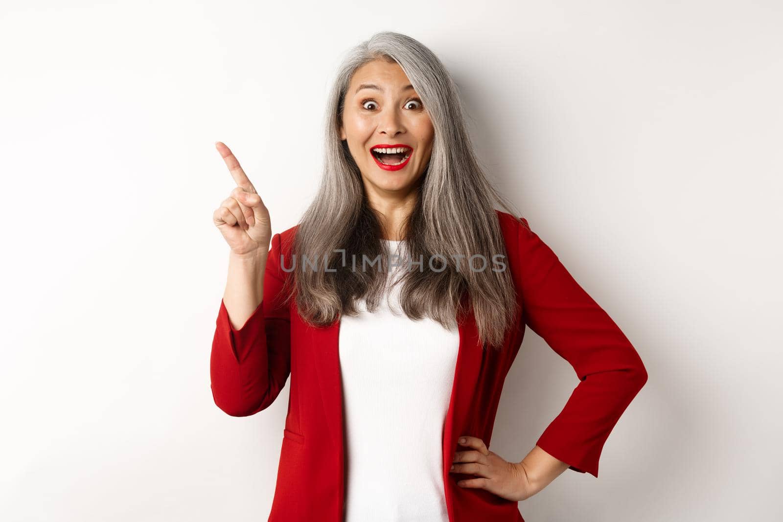 Business concept. Cheerful asian businesswoman with grey hair, wearing red blazer and makeup, pointing upper left corner and smiling amazed, white background.