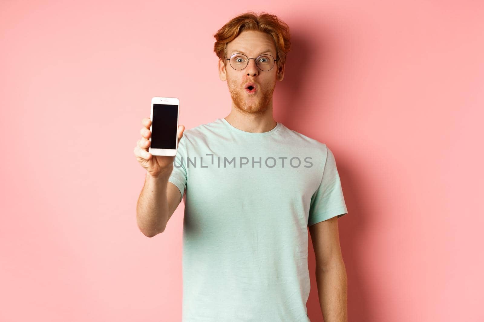 Image of handsome guy with red hair, wearing glasses and t-shirt, saying wow and showing smartphone screen, standing against pink background by Benzoix