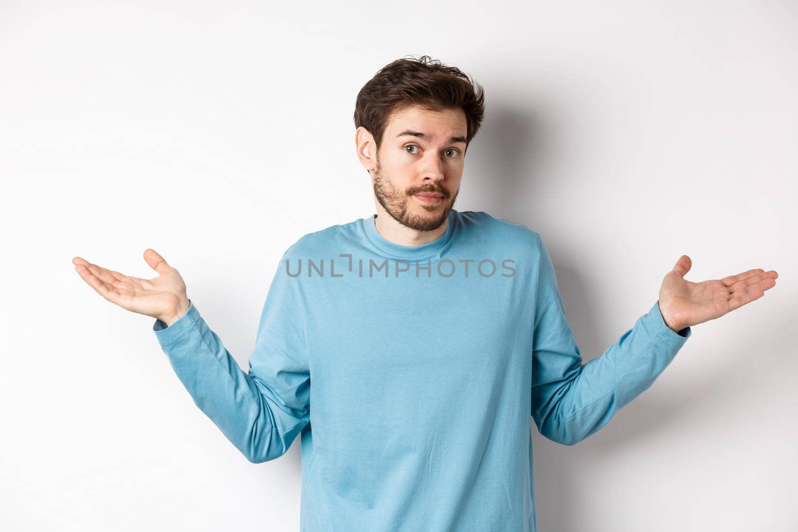 Clueless bearded guy in blue shirt, shrugging shoulders and looking unaware at camera, know nothing, standing on white background.