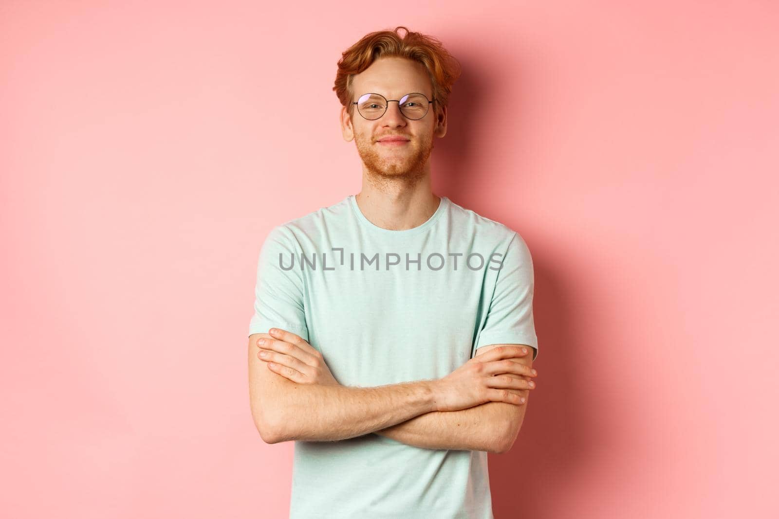 Portrait of satisfied caucasian man with red hair and beard, cross arms on chest and smiling with smug face, wearing glasses, standing over pink background.