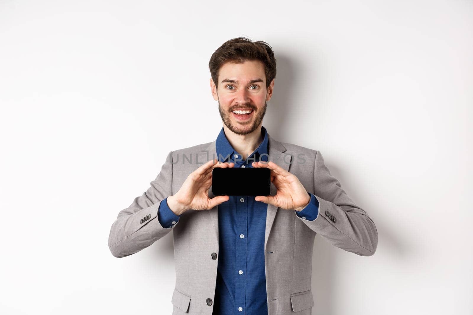 E-commerce and online shopping concept. Excited young businessman in suit showing empty smartphone, screen demonstrate app achievement, white background.