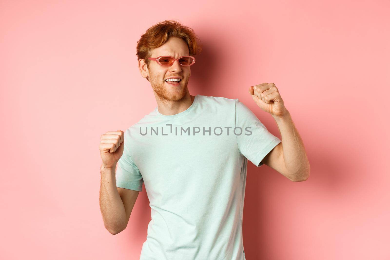 Tourism and vacation concept. Cheerful redhead man having fun at party, dancing and enjoying holiday, standing in sunglasses and t-shirt against pink background.