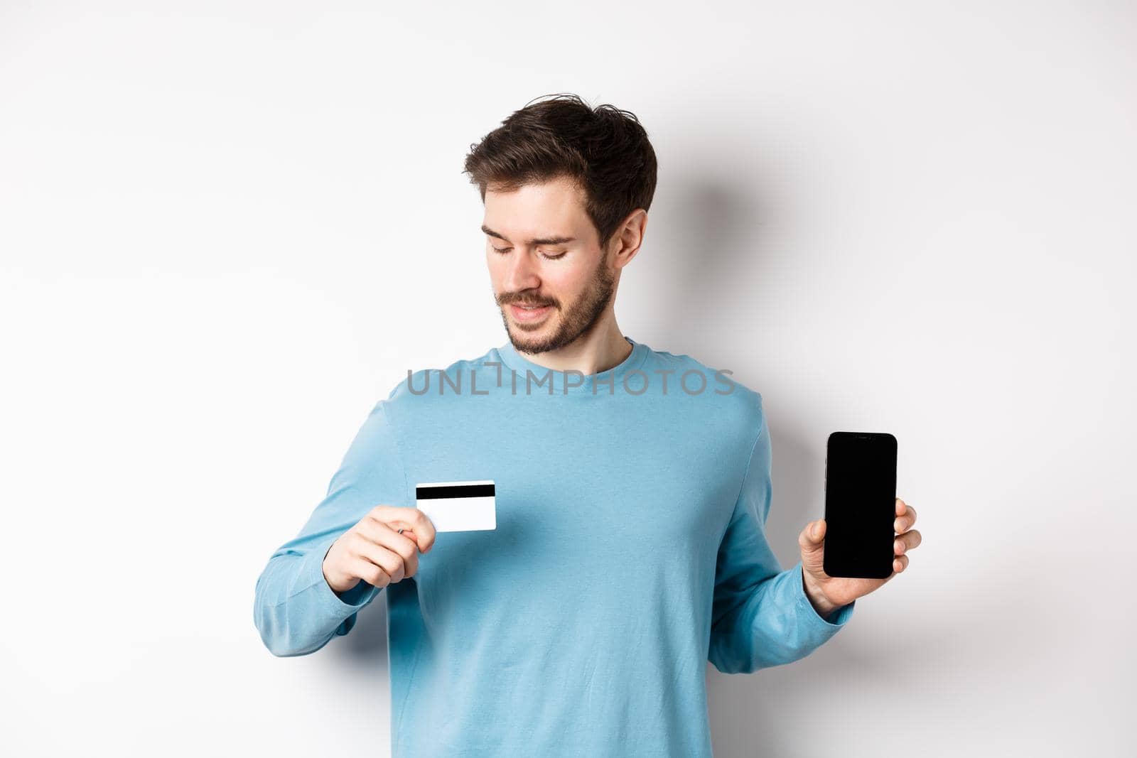 Handsome young man with beard, showing empty smartphone screen and looking at plastic credit card, standing over white background by Benzoix