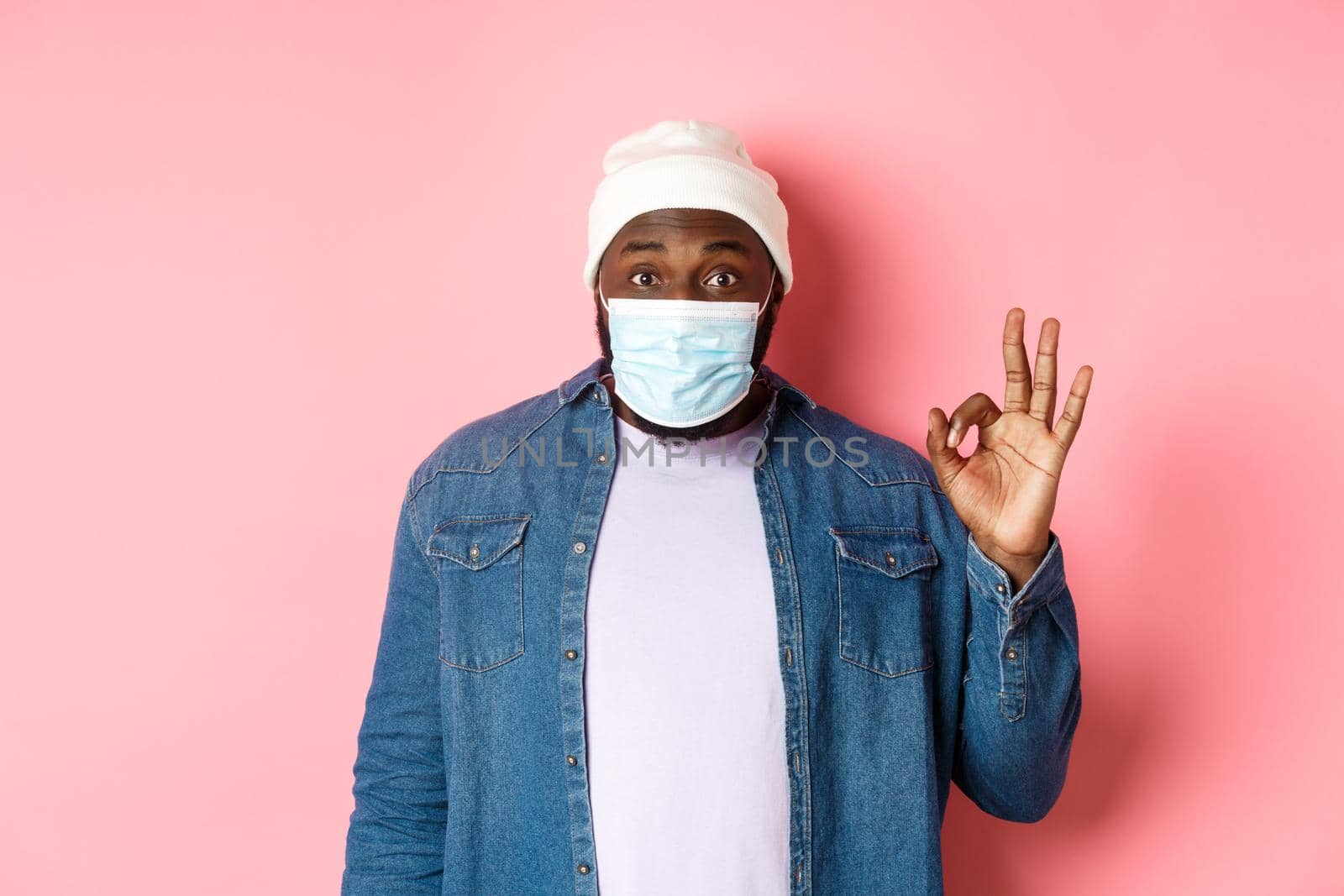 Coronavirus, lifestyle and social distancing concept. Impressed african-american man in face mask, showing okay sign, like and agree, standing over pink background.