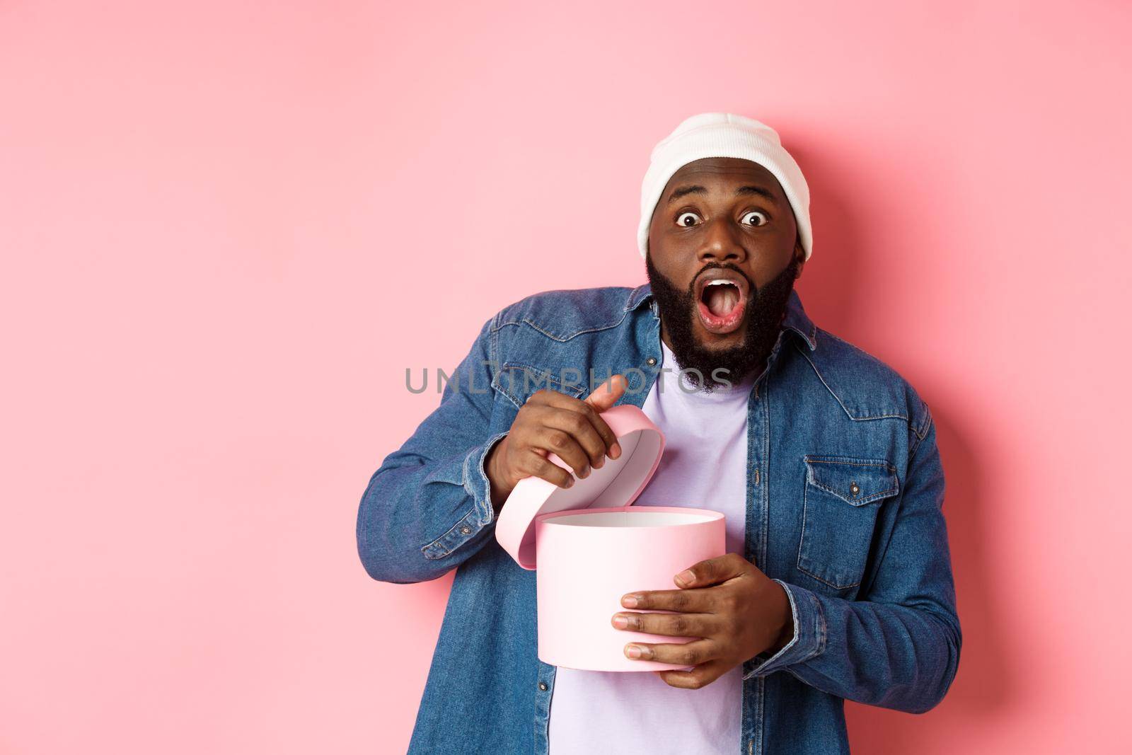 Excited Black man open box with gift and staring at camera grateful and amazed, receiving present, standing over pink background by Benzoix