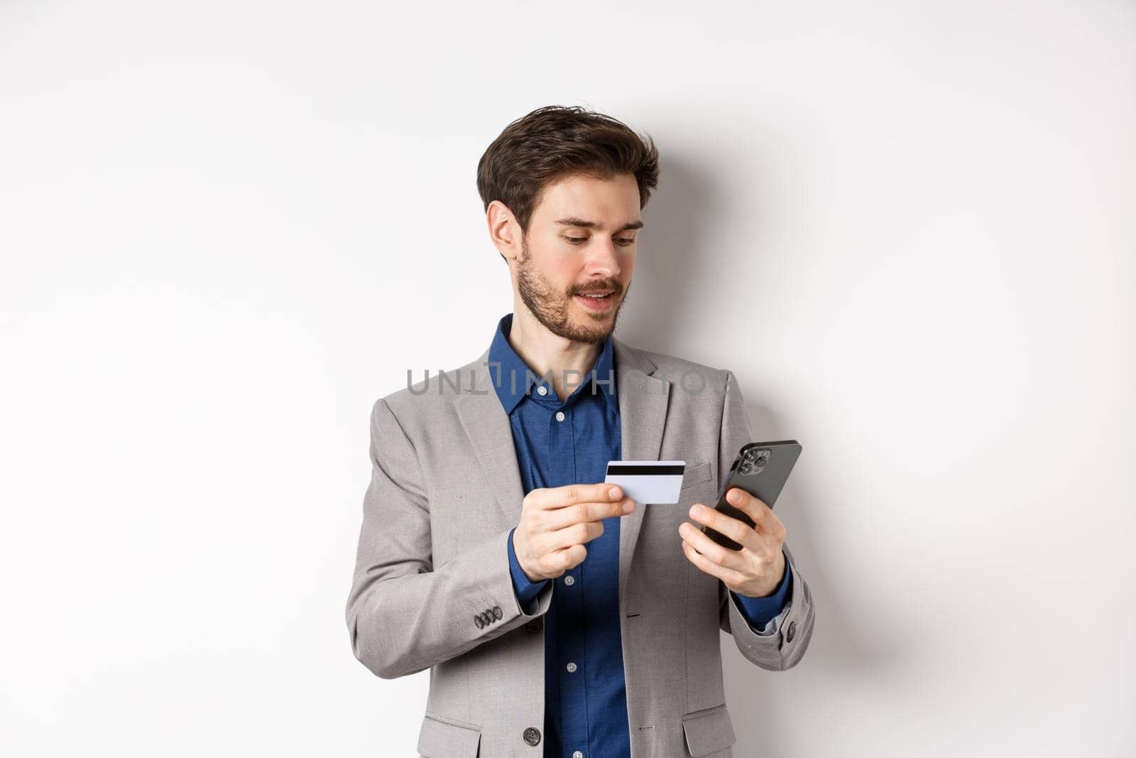 Online shopping. Smiling businessman paying with credit card on smartphone, sending money, standing in suit on white background.