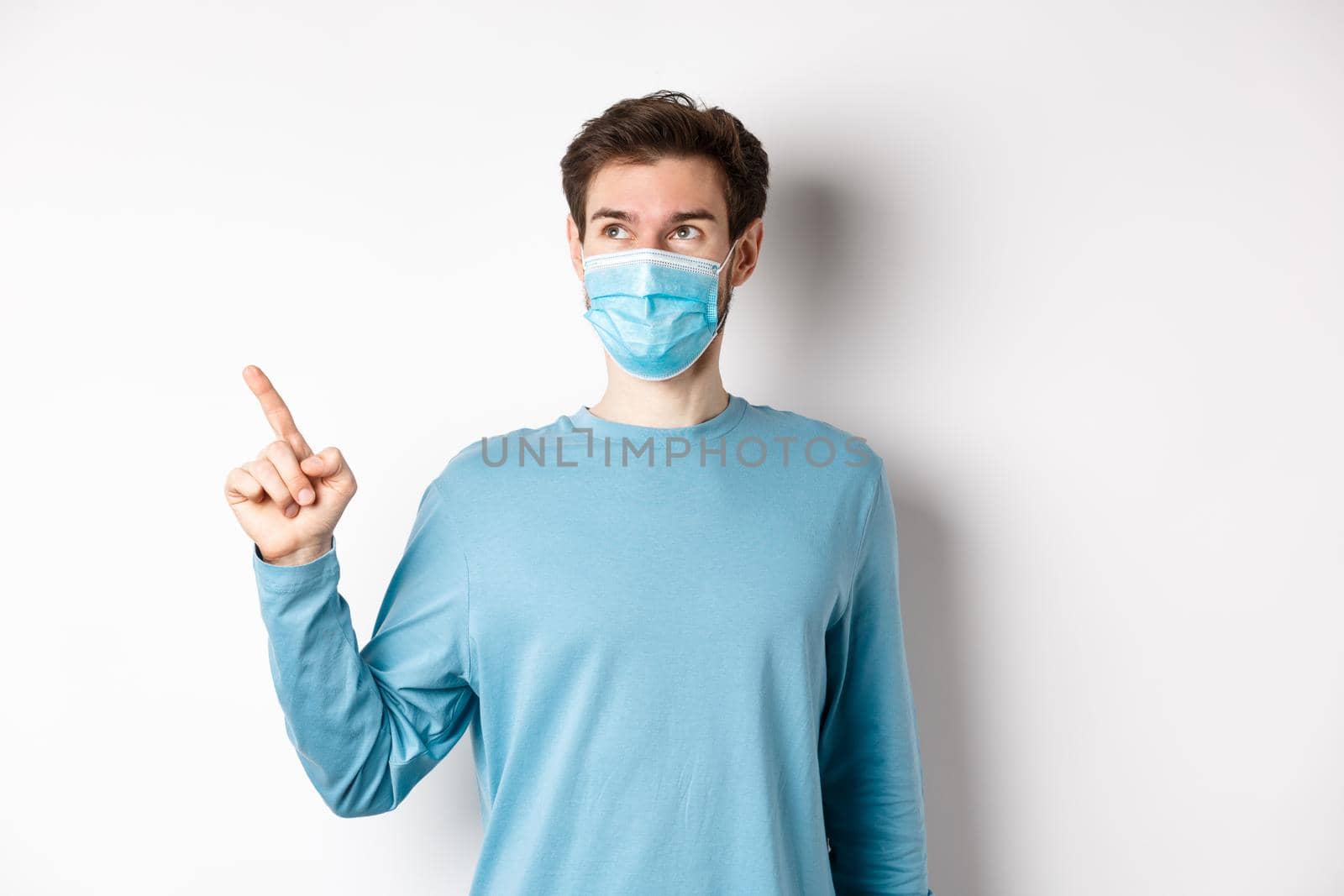 Coronavirus, health and quarantine concept. Man in medical mask smiling pleased, looking and pointing finger left at logo, showing promo offer, white background.