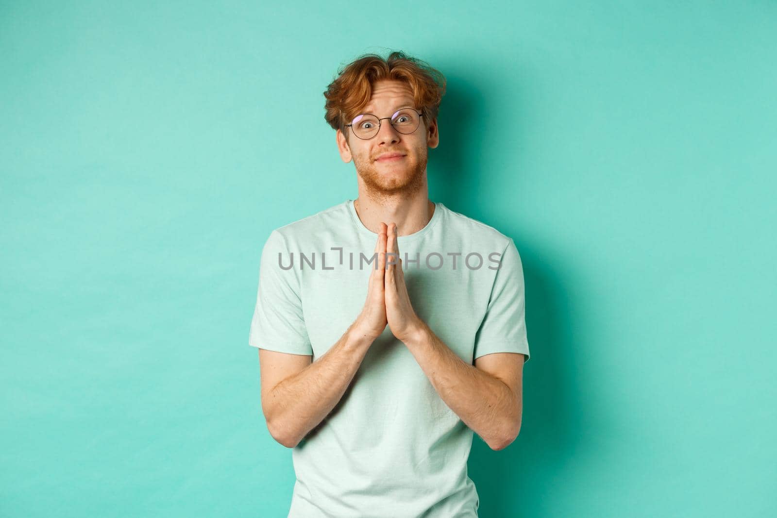 Cute redhead man asking for help, saying please with namaste gesture, pleading or begging for favour, looking silly at camera, standing over mint background.