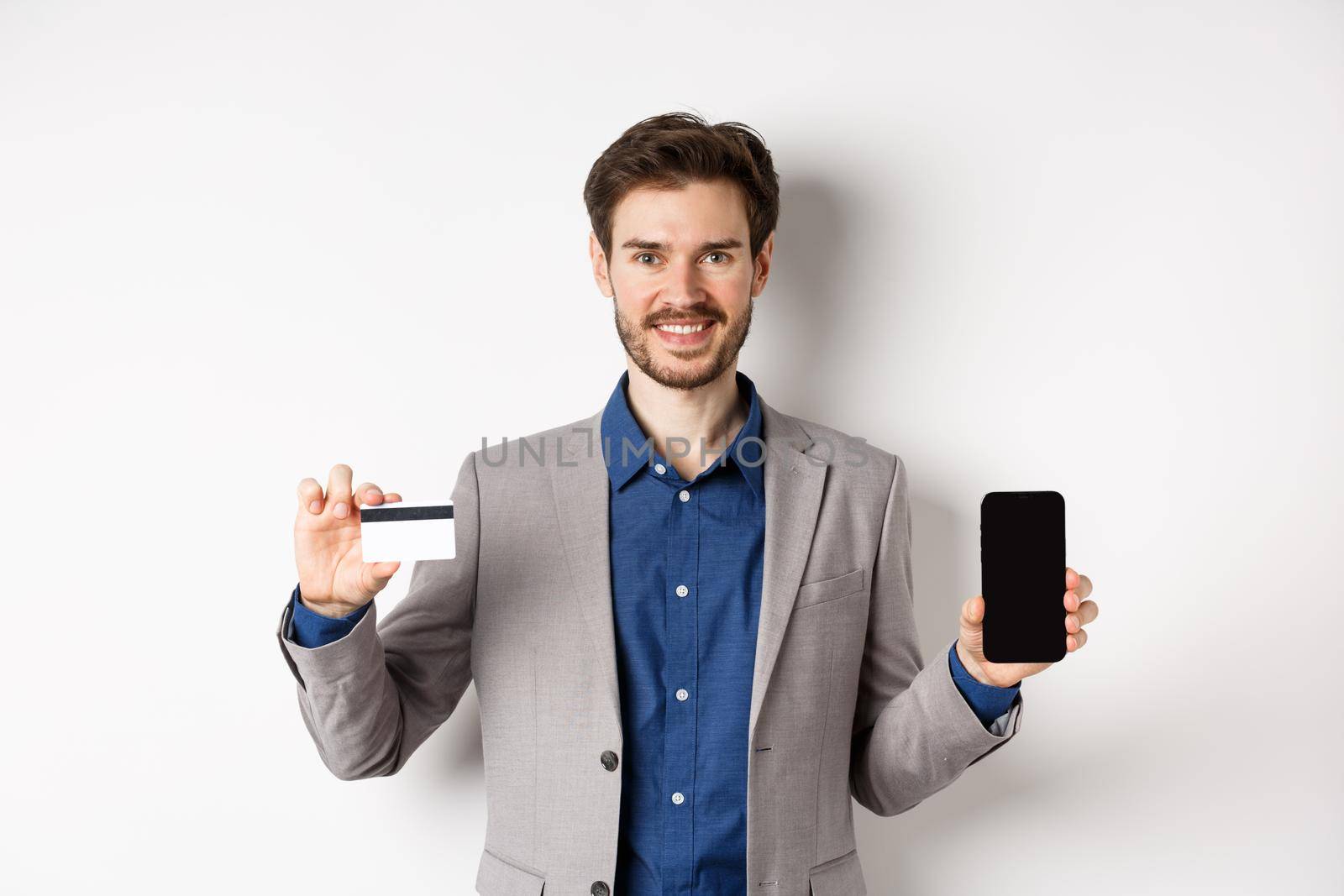 Online shopping. Smiling business man in suit showing plastic credit card with empty smartphone screen, standing against white background by Benzoix