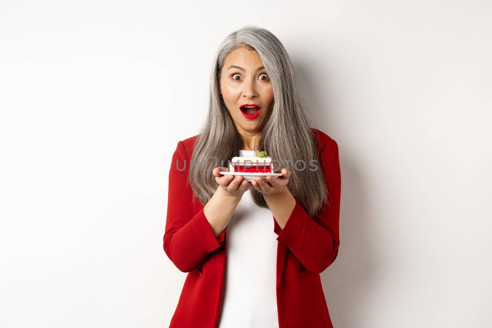 Surprised asian middle-aged woman holding piece of tasty cake, looking amazed at camera, standing over white background.