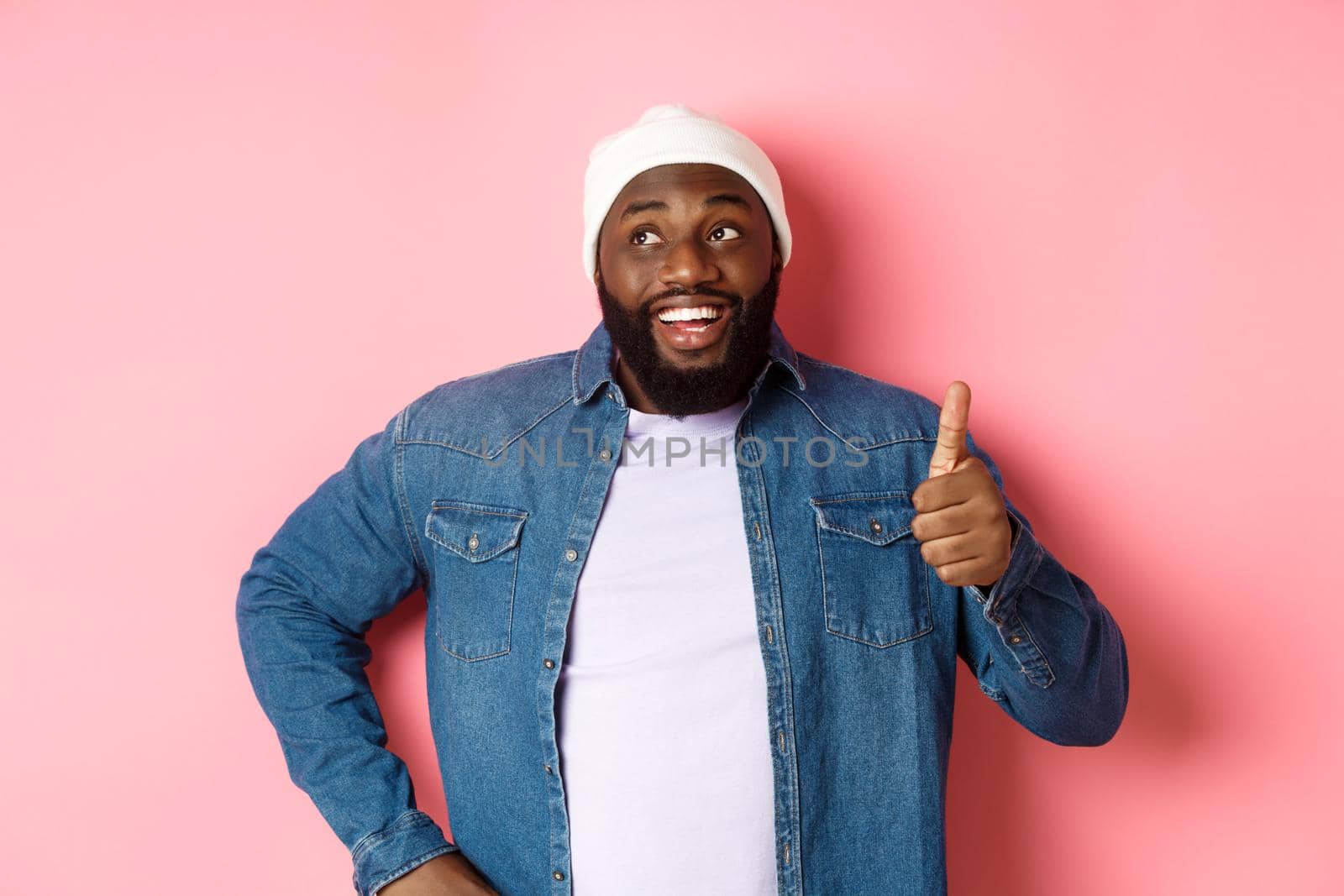 Happy Black man showing thumb-up, looking at upper left corner thoughtful, standing over pink background by Benzoix