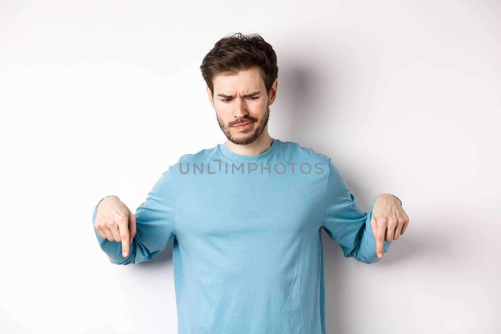 Skeptical boy pointing fingers down and frowning disappointed, looking at bad promo offer, standing on white background.