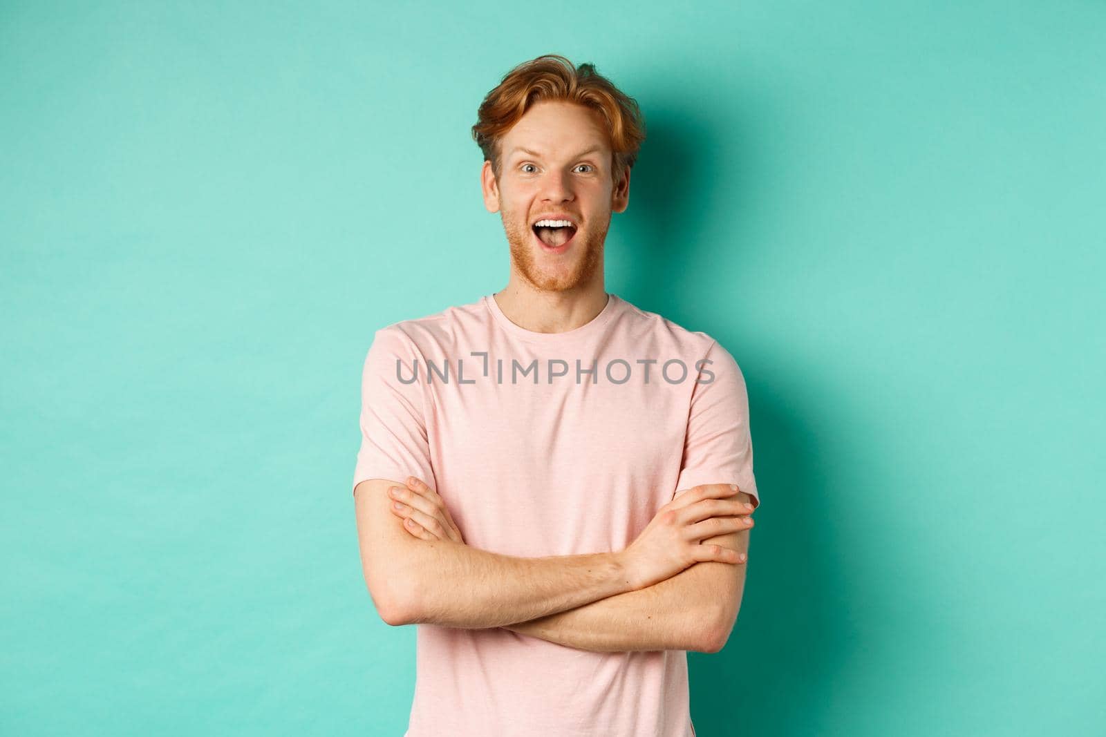Enthusiastic redhead man in t-shirt checking out interesting promo, cross arms on chest and looking at camera with awe, standing over turquoise background by Benzoix