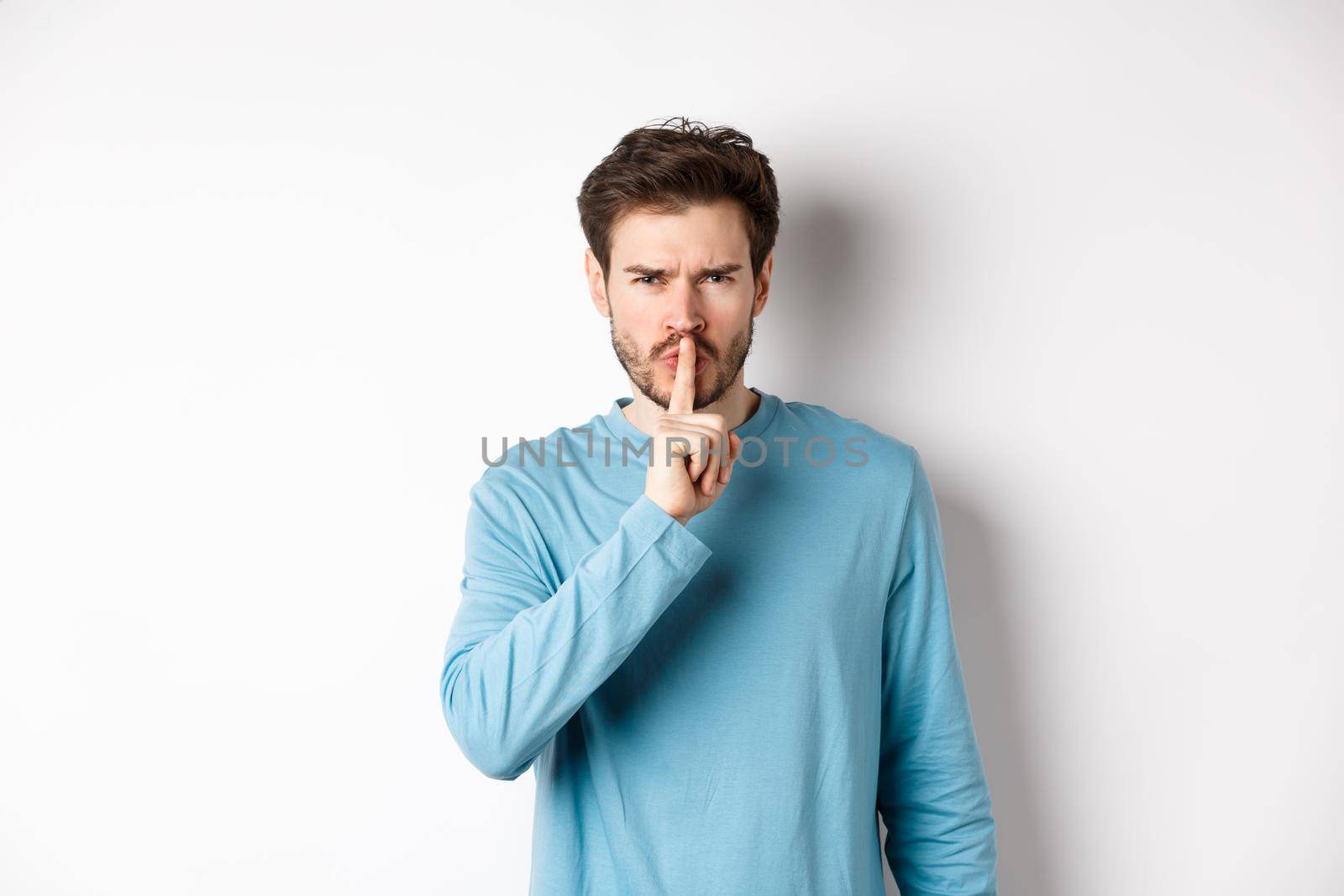 Angry man shushing at camera and telling to be quiet, frowning upset, standing over white background with finger on lips, taboo gesture.
