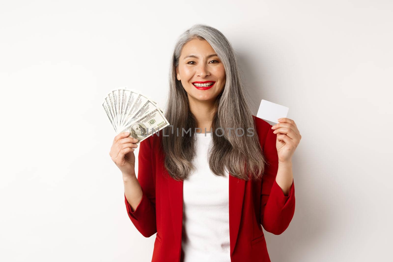 Successful asian senior businesswoman showing money in dollars and plastic card, smiling happy at camera, wearing red blazer and make-up.