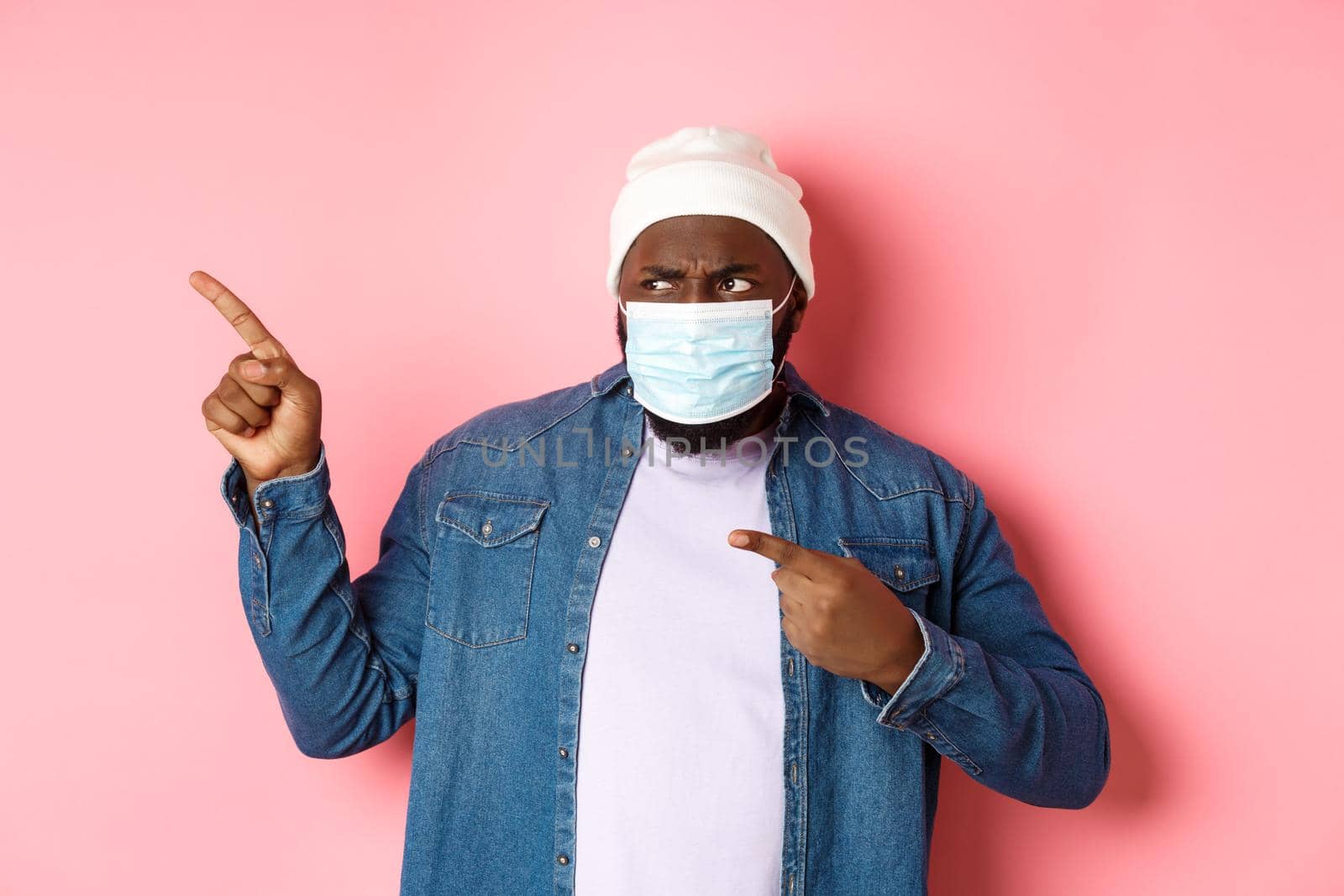 Coronavirus, lifestyle and global pandemic concept. Confused and disappointed african-american guy in face mask pointing fingers left, staring upset with camera, pink background.