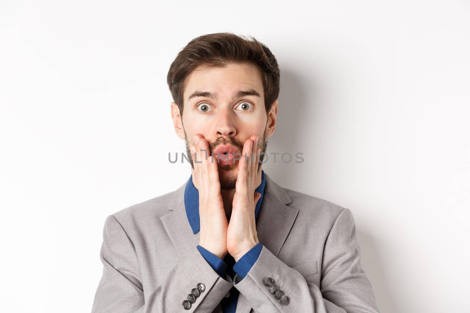 Surprised businessman in suit look with disbelied, pucker lips and touching face as staring at camera, standing on white background.