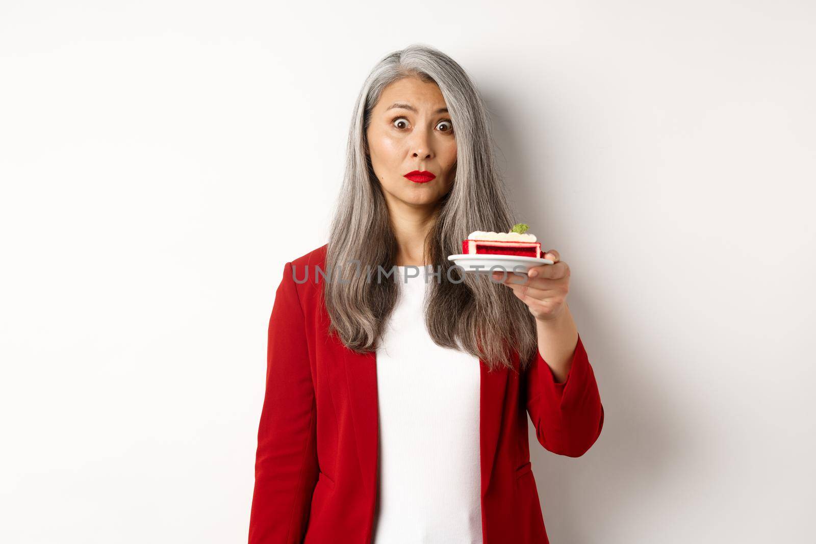 Portrait of mature asian woman on diet holding sweet cafe, looking indecisive at camera, standing over white background.