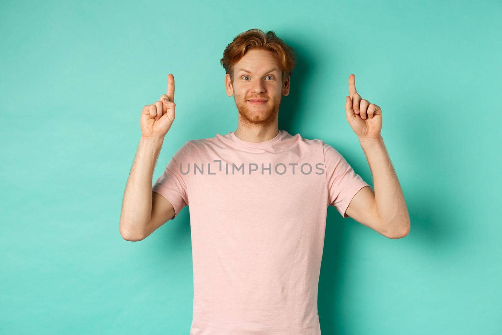 Attractive bearded man with red hair, wearing t-shirt, smiling cheerful and pointing fingers up, showing advertisement, standing over turquoise background.