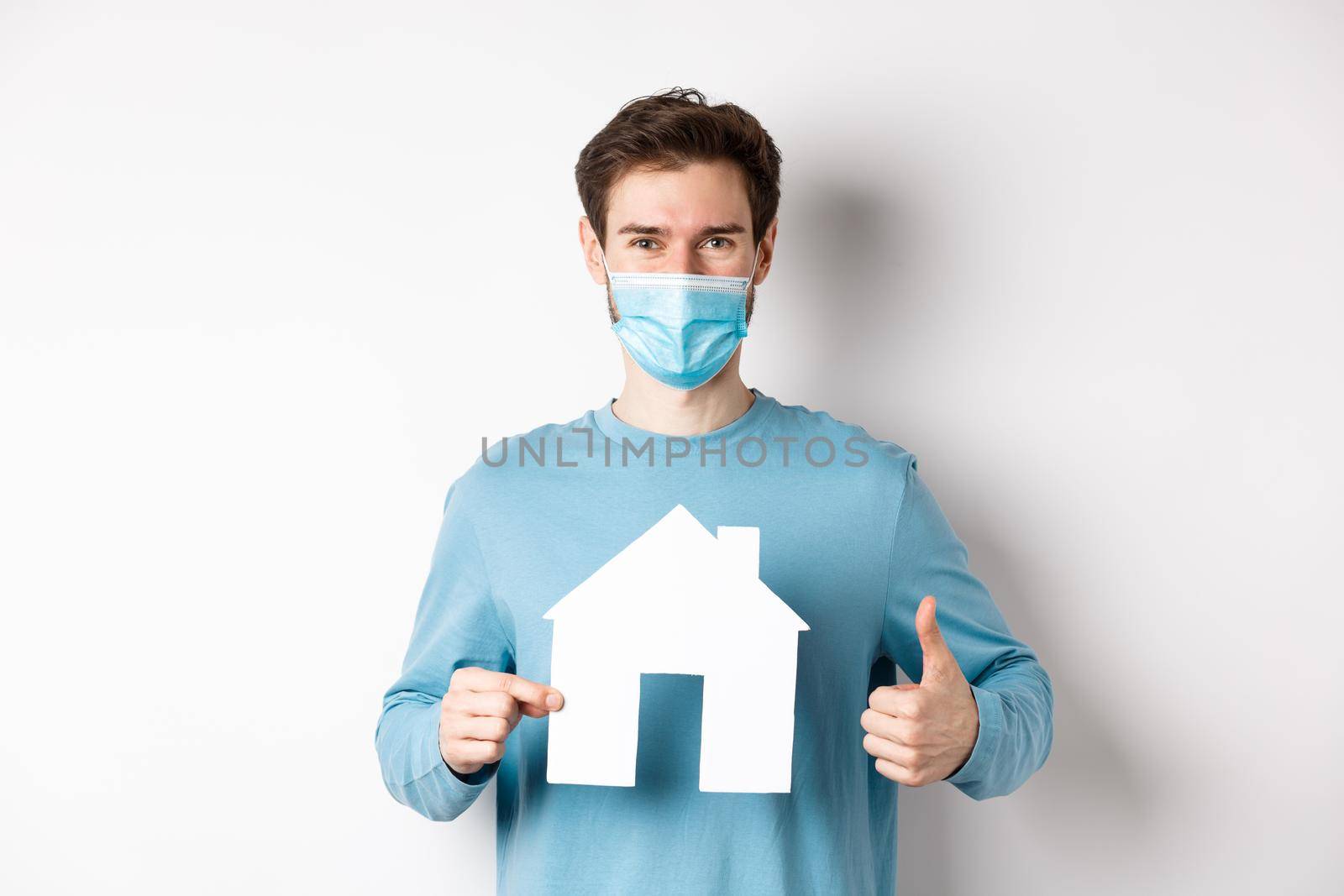 Covid and real estate concept. Smiling agency client showing thumb up and paper house cutout, wearing medical mask, approve good deal by Benzoix