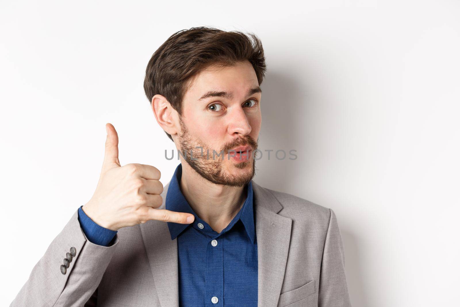 Handsome macho guy in suit showing phone sign near ear, flirting and asking to call him, giving his number, white background.