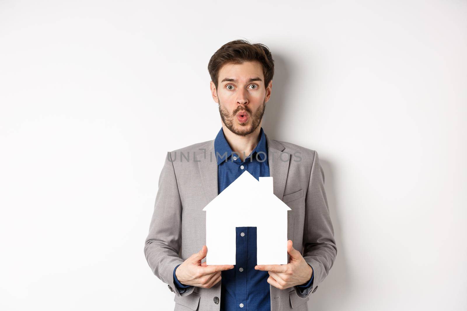 Real estate. Amazed caucasian man in suit checking out special offer for property, standing with paper house cutout and say wow at camera, white background.