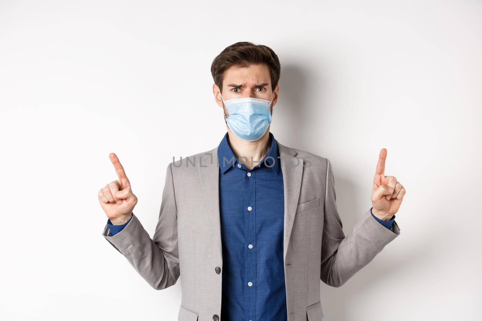 Covid-19, pandemic and business concept. Disappointed frowning guy in suit and medical mask pointing fingers up, complaining on unfair situation, white background by Benzoix