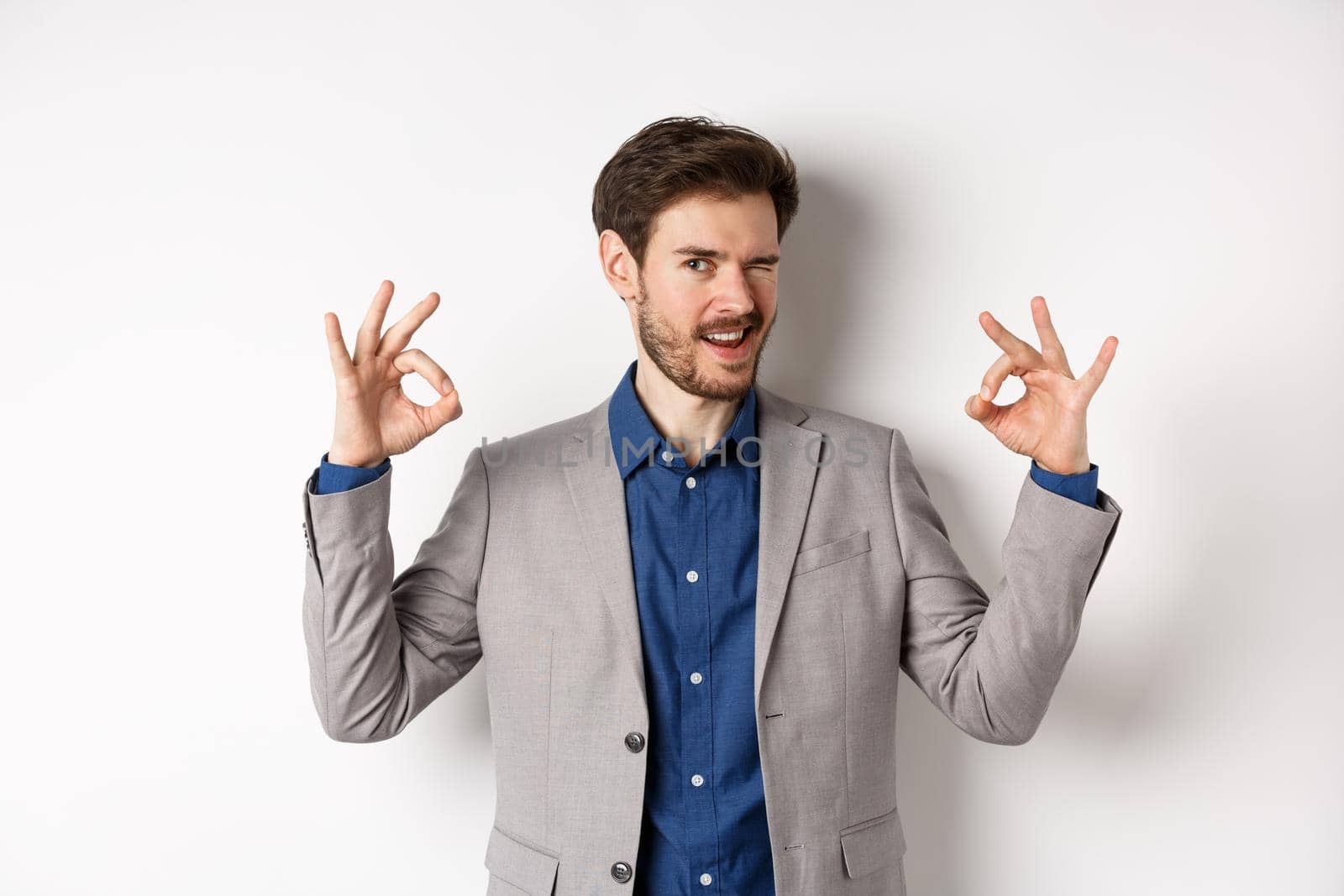 Cheeky handsome man in suit winking at you, showing okay signs, all under control gesture, approve good choice, assuring everything OK, standing on white background.