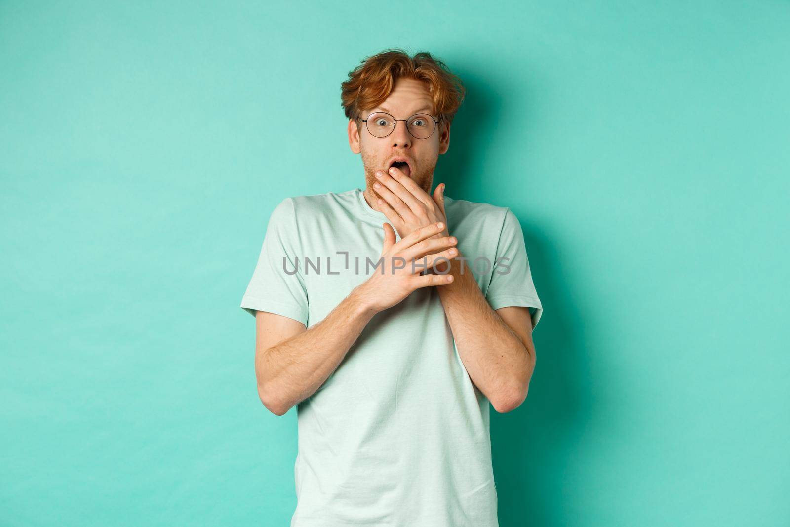 Shocked redhead man in glasses gasping startled, covering mouth and staring at camera scared, standing over turquoise background by Benzoix