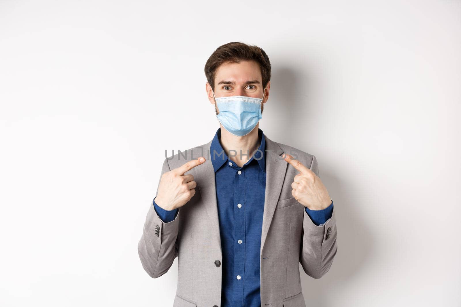 Covid-19, pandemic and business concept. Excited businessman in suit pointing at his medical mask and smiling with eyes, white background.