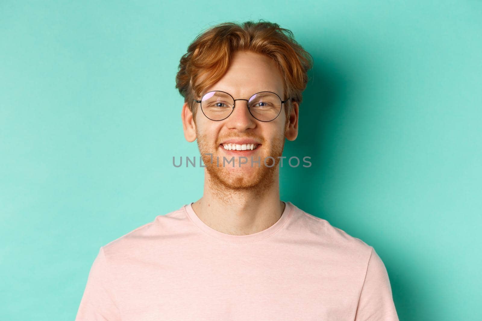 Close up of handsome redhead man in glasses looking at camera, smiling with white teeth, standing against mint background.