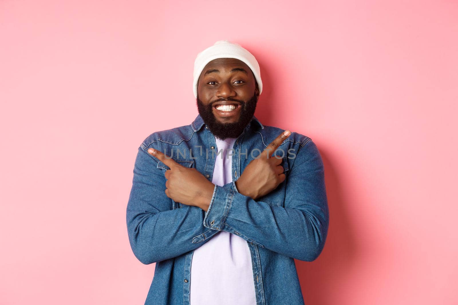 Happy young african-american hipster guy pointing fingers sideways, smiling and showing two choices, showing offers, standing over pink background.