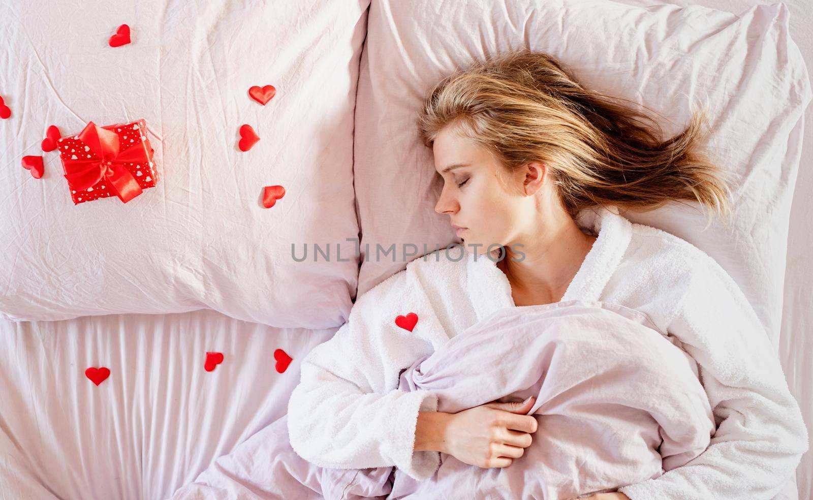 Happy valentines day. young blond woman sleeping in bed with holiday gift and red confetti