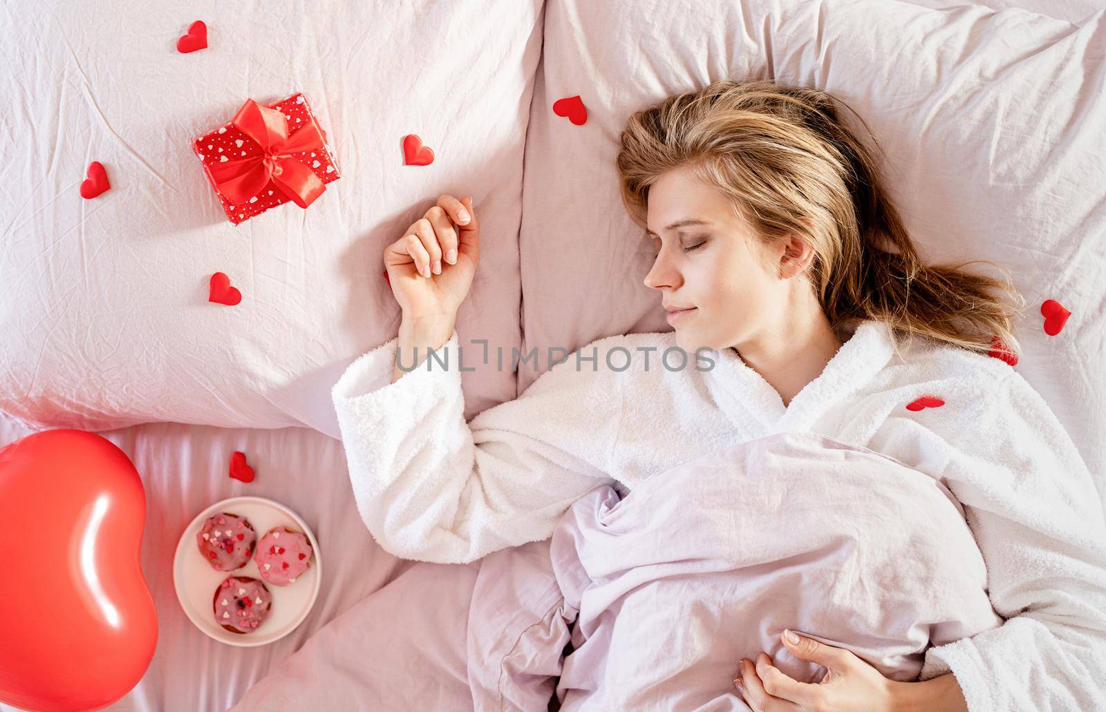 young blond woman lying in bed with holiday gift and red confetti by Desperada