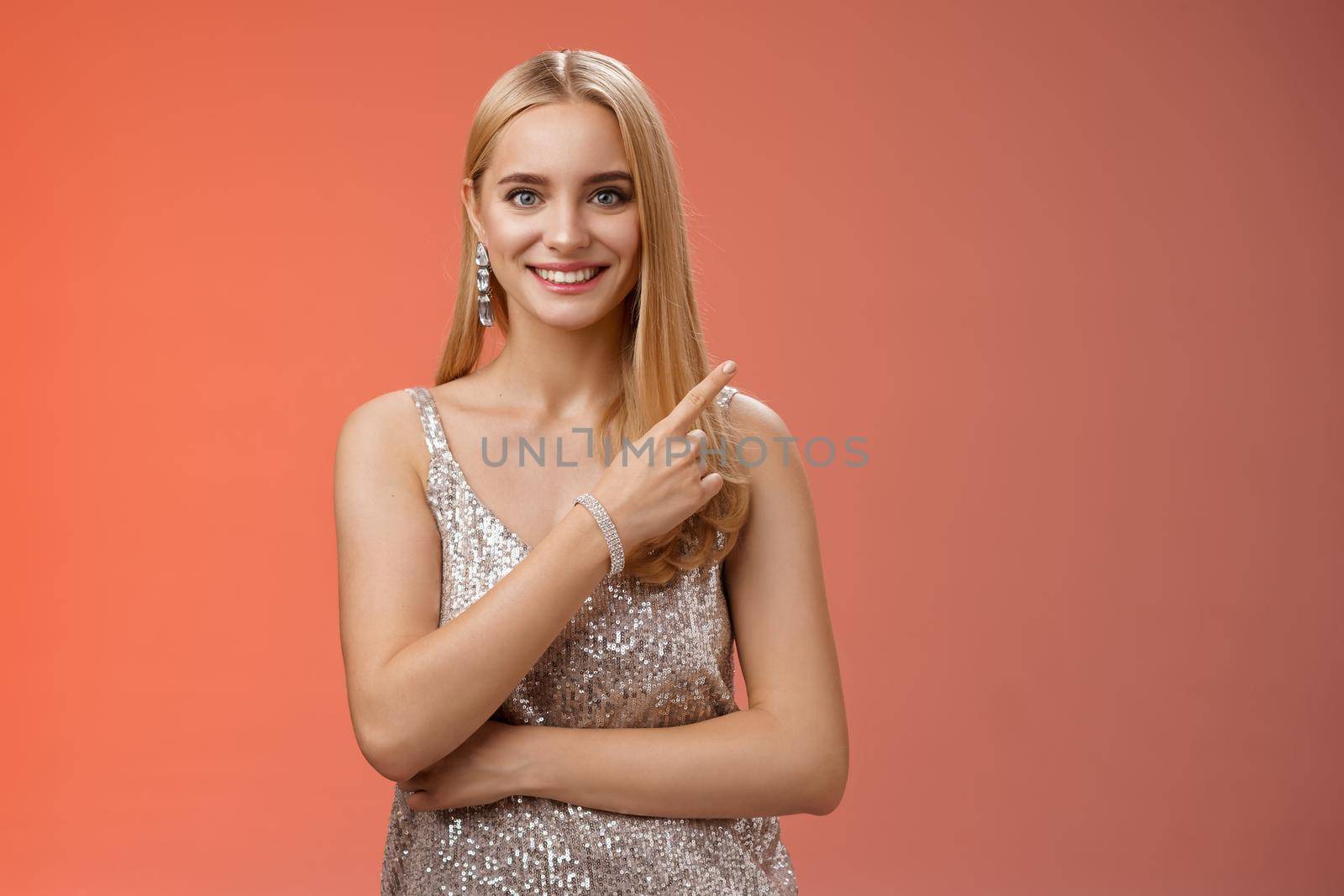 Confident attractive feminine glamour blond young woman in silver glittering dress smiling broadly pointing up discuss recent news promo sales standing friendly grinning camera red background by Benzoix