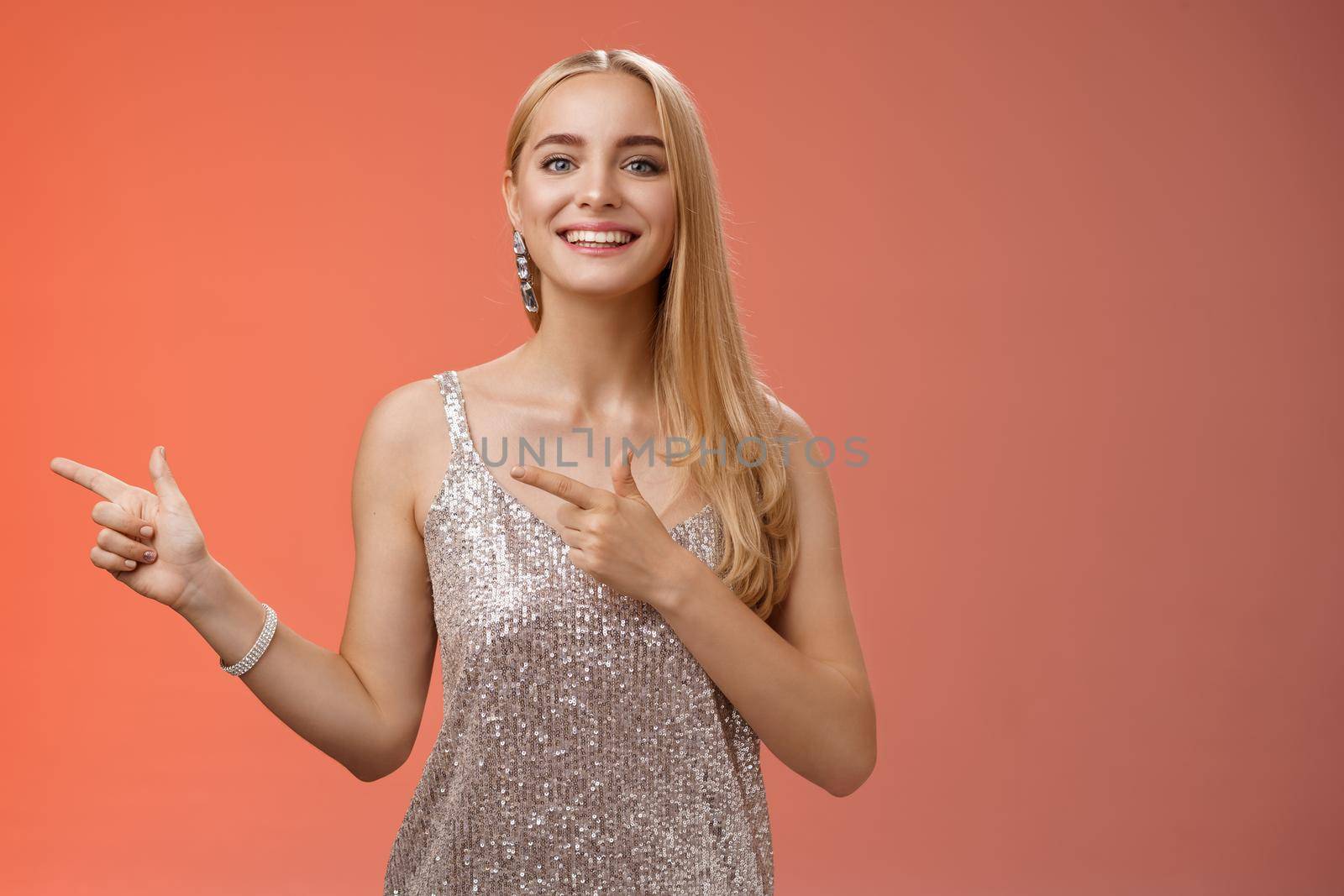Proud happy good-looking stylish blond attractive caucasian woman in silver luxurious glamour dress pointing left smiling broadly showing friends new apartment, standing red background.