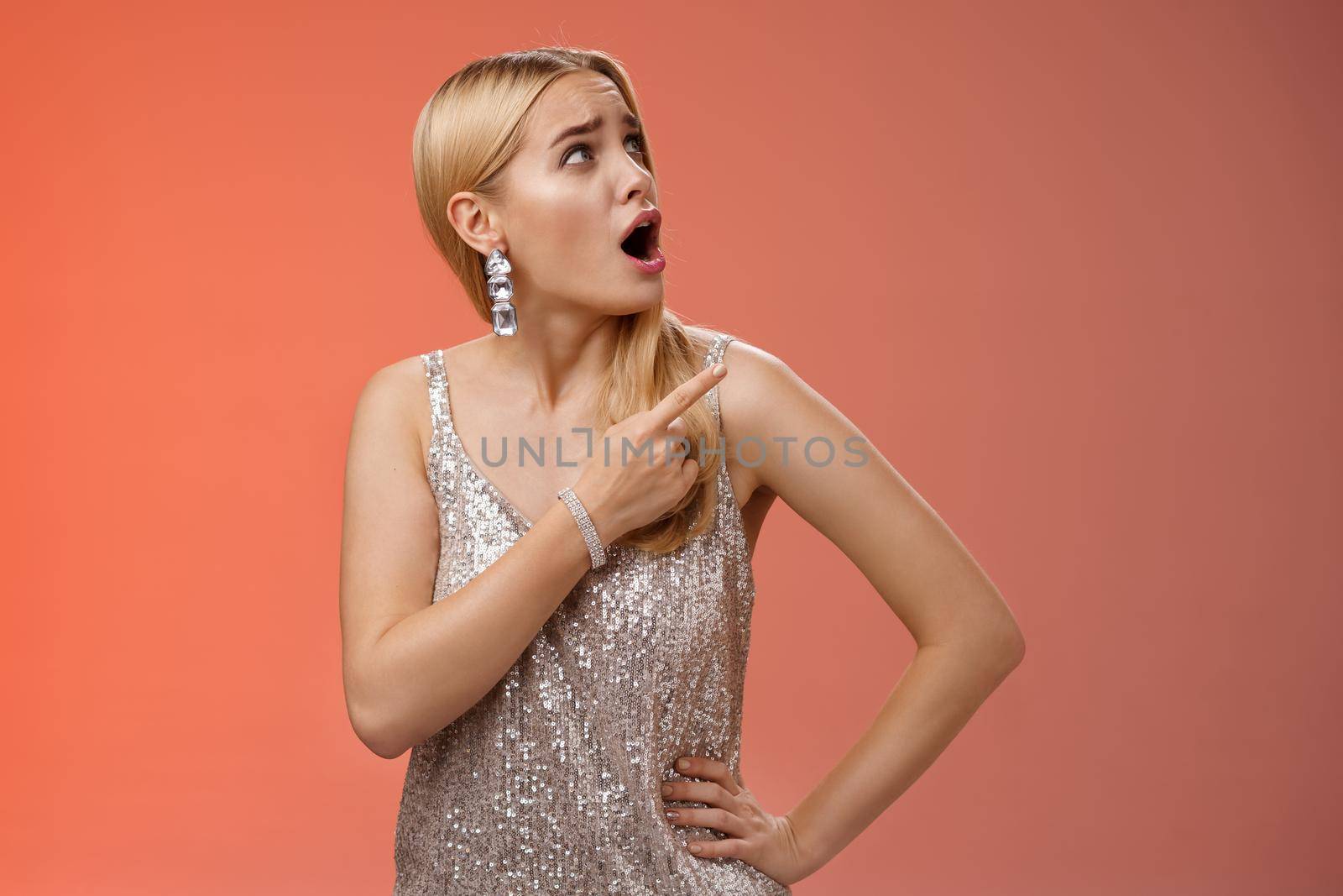 Shocked displeased bothered arrogant blond glamour woman in silver glittering dress turning upper right corner pointing complaining strange noise come upstairs, standing questioned red background.