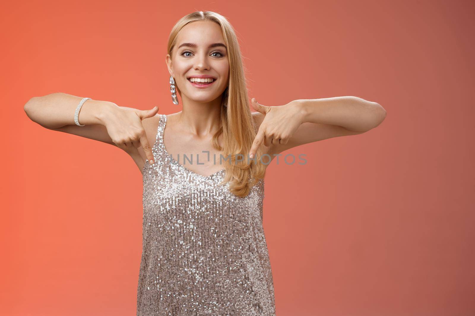 Attractive glamour blond woman in silver glittering dress pointing down smiling excited showing awesome party place hang-out inviting try-out standing pleased red background enjoying perfect evening.