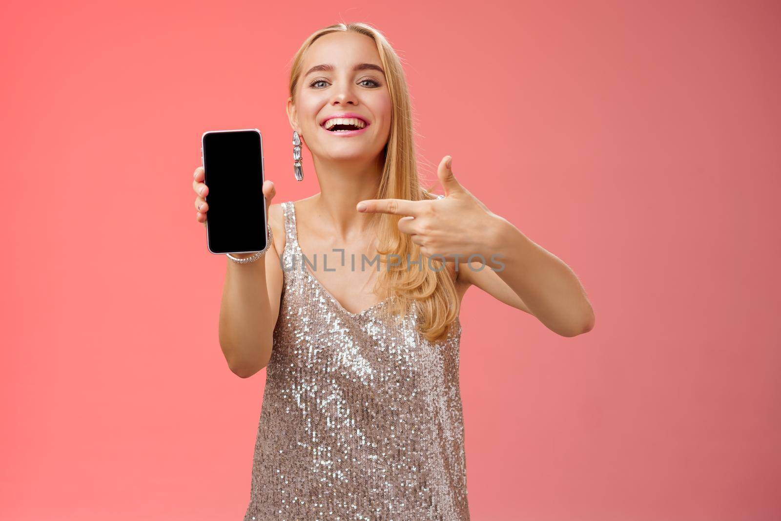 Boastful proud charming elegant blond woman in stylish evening dress show smartphone display proudly pointing mobile phone screen smiling showing photo boyfriend, standing red background by Benzoix