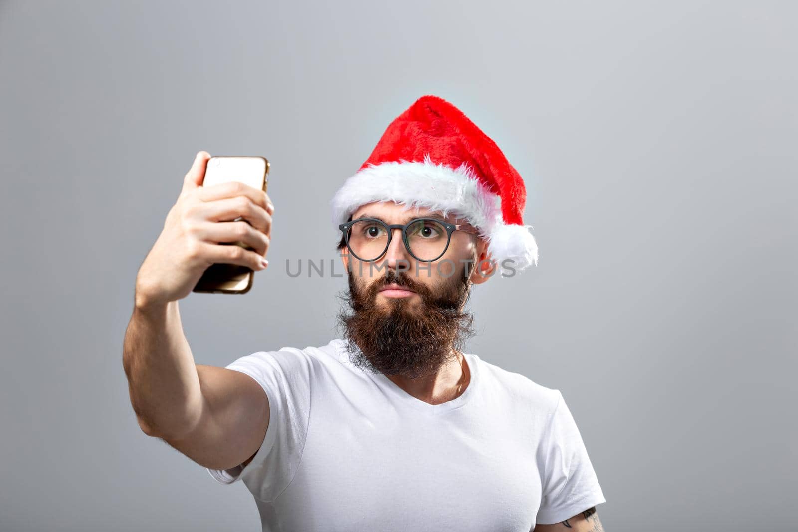 Christmas, holidays, technology and people concept - handsome bearded man in santa hat taking selfie picture with smartphone on grey background.