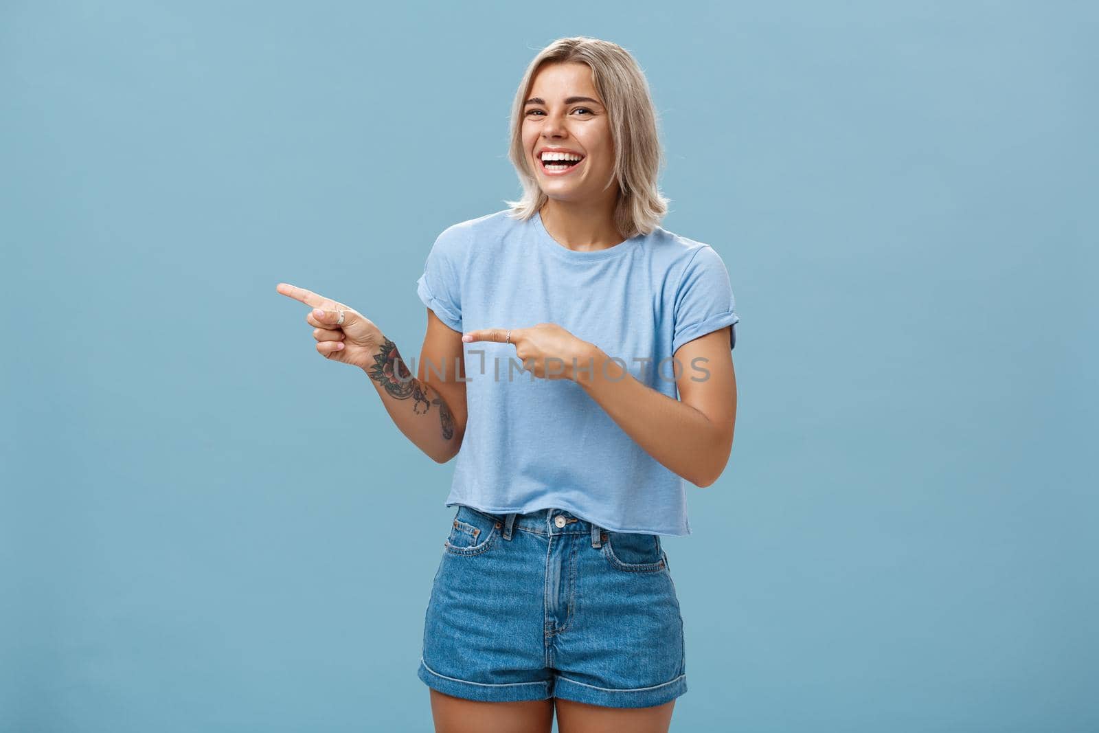 Charismatic joyful attractive athletic woman with fair hair and tanned skin laughing out loud with amusement pointing left and gazing at camera happily standing entertained over blue wall. Copy space