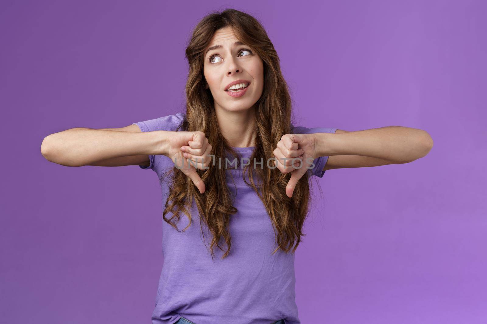 Unimpressed ignorant snobbish attractive curly-haired girl disagree lame idea look away disappointed show thumbs down dislike disapproval gesture not interested upset purple background.