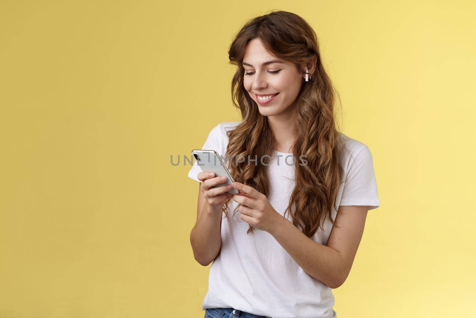Happy caring tender feminine girlfriend curly long hair hold smartphone picking song listen way home smiling broadly look mobile phone scroll music platform wear wireless earbud yellow background. Lifestyle.