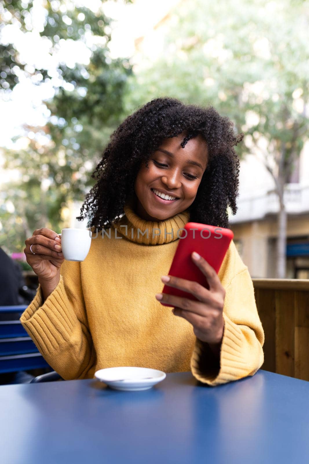Happy young African American woman using mobile phone drinks coffee in an outdoors coffee shop terrace. Vertical image. by Hoverstock