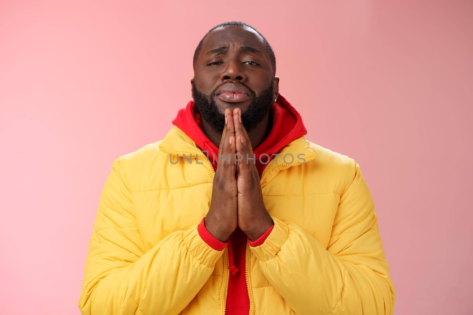 Miserable cute african-american bearded man in yellow coat asking help begging press palms supplicating apologizing please help, standing pink background sad need advice lending money.