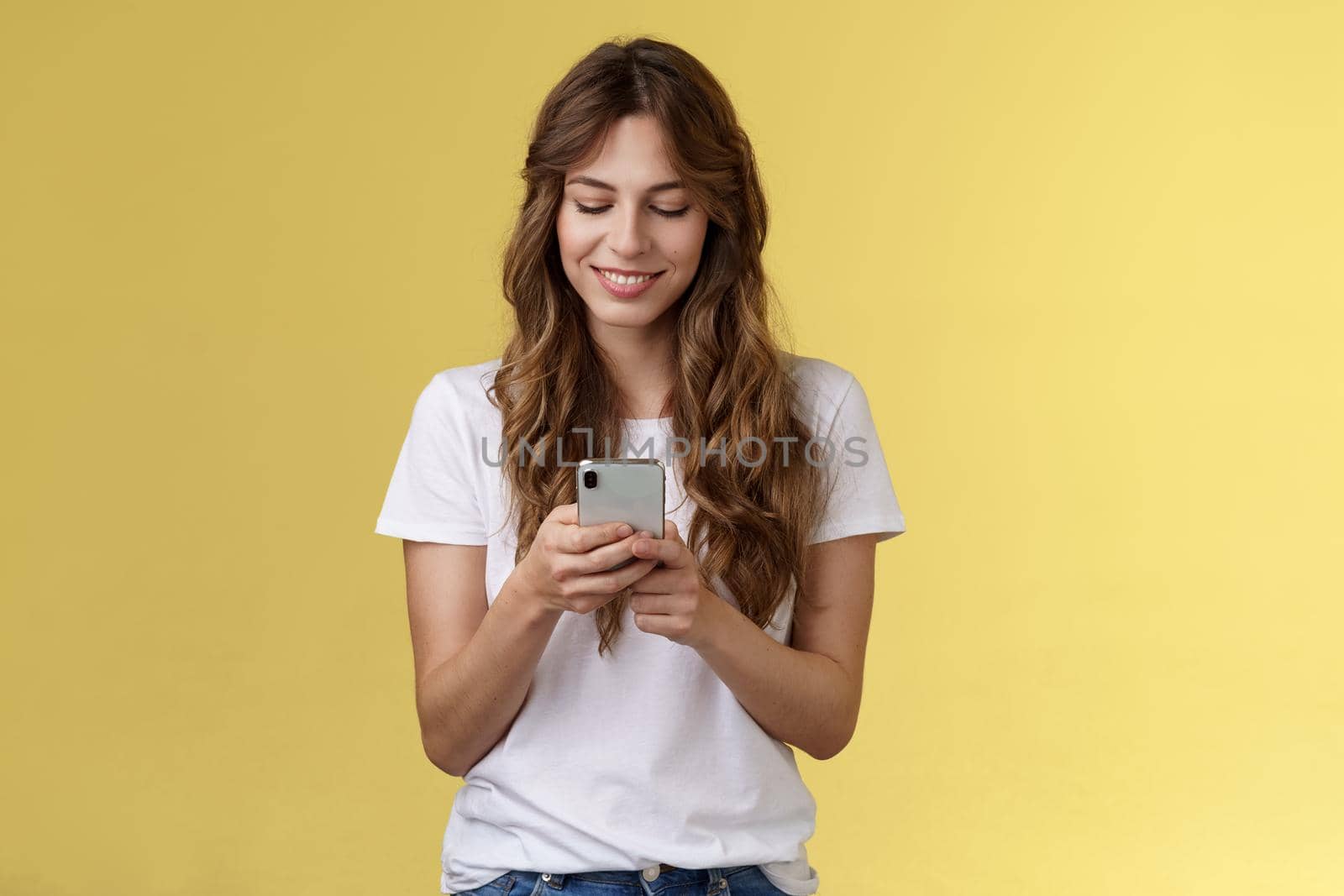 Lovely feminine tender caucasian girl white t-shirt jeans hold smartphone texting messaging girlfriend smiling delighted look tenderly lovely smile mobile phone screen yellow background.
