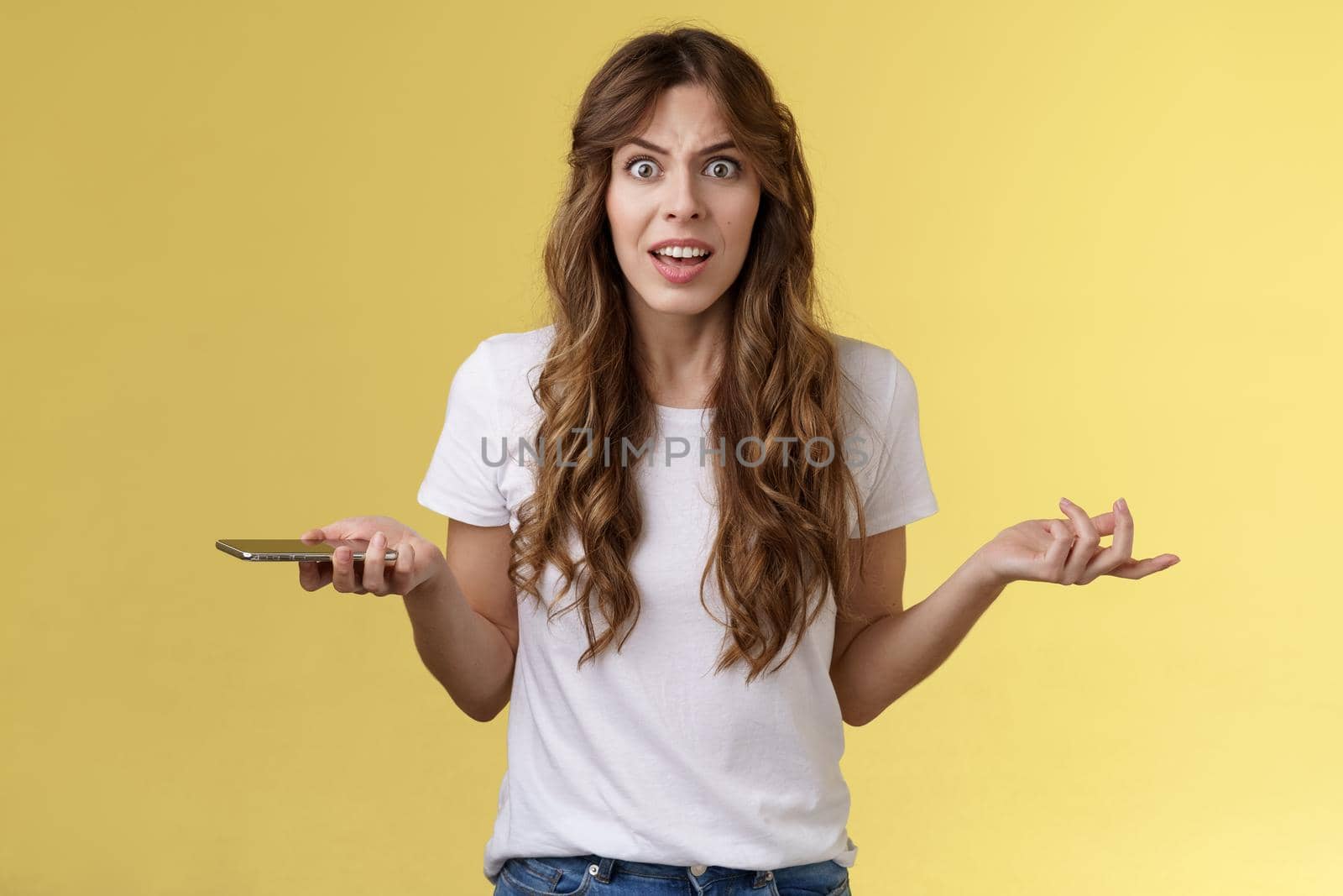 Pissed frustrated bothered curly-haired caucasian girl stare camera puzzled complain hold smartphone shrugging spread hands sideways dismay arguing after intense serious phonecall yellow background.