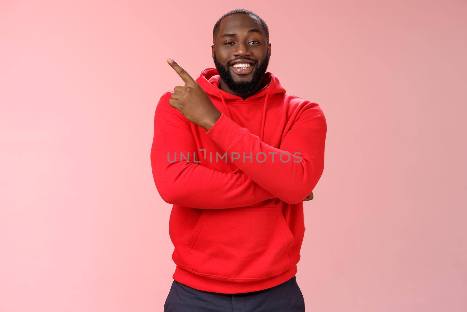 Cheeky outgoing smiling attractive african-american male model in red hoodie grinning energized pointing upper left corner indicating awesome product recommend promo, standing pink background.